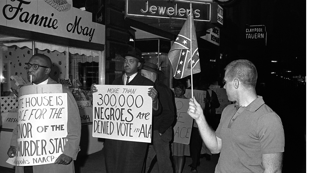 A man holds a Confederate flag as demonstrators, including one carrying a sign saying 'More than 300,000 Negroes are Denied Vote in Ala', demonstrate in front of an Indianapolis hotel where then-Alabama Governor George Wallace was staying [Bob Daugherty/AP/File]