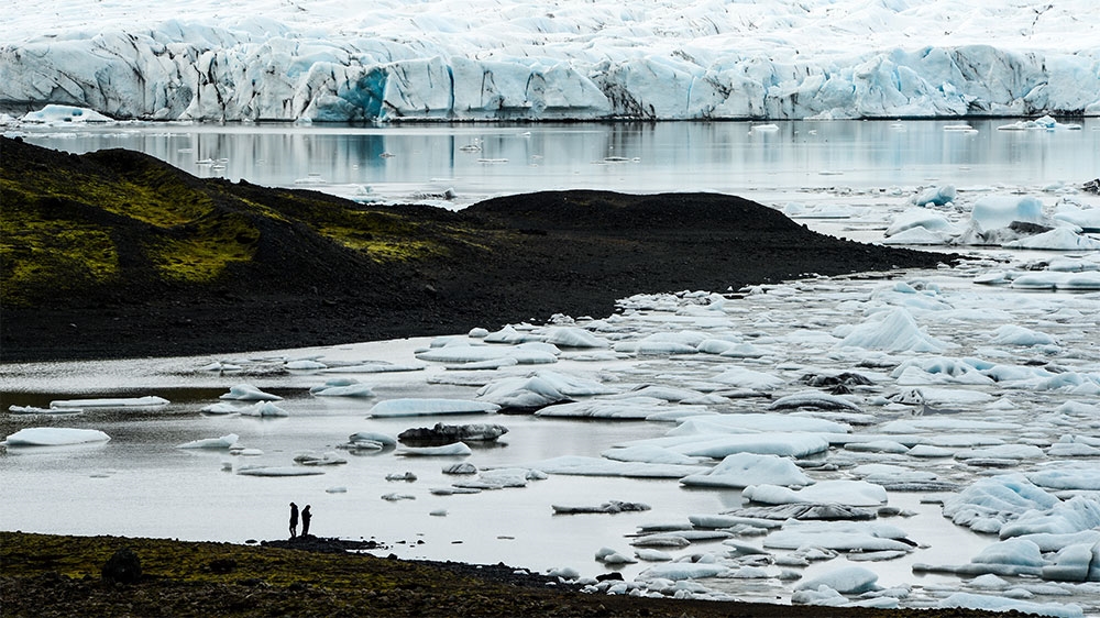 Two glacioloigsts taking temperature readings stand at the edge of the Fjallsarlon glacial lagoon. Large icebergs breaking off from the tongue float to the edge of the lagoon and eventually drift out into the sea [Alexander Lerche/Al Jazeera]