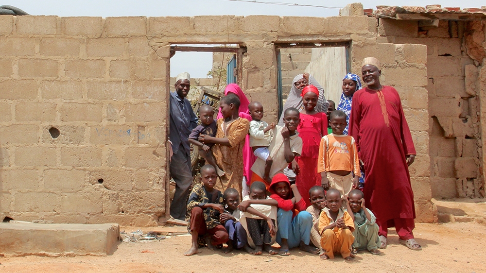 Sani Baga, far right, his daughter Zulai (wearing a black-and-white striped hijab) and their family stand in front of the compound in Kano that Haruna Ya’u (standing in the doorway) divided in two to accommodate a family in need [Femke van Zeijl/Al Jazeera]