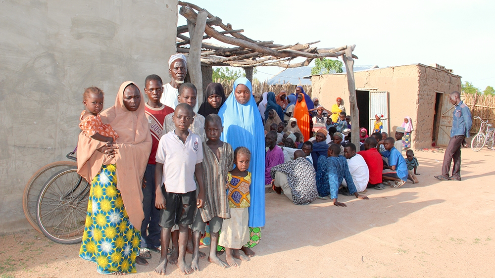 Mohammed Jidda stands in front of his compound with his first wife Fatima (who wears a black hijab) and his second wife Mairo, far left, whom he married in 2014, and his children. Behind them sit some of the people hosted by the village [Femke van Zeijl/Al Jazeera]