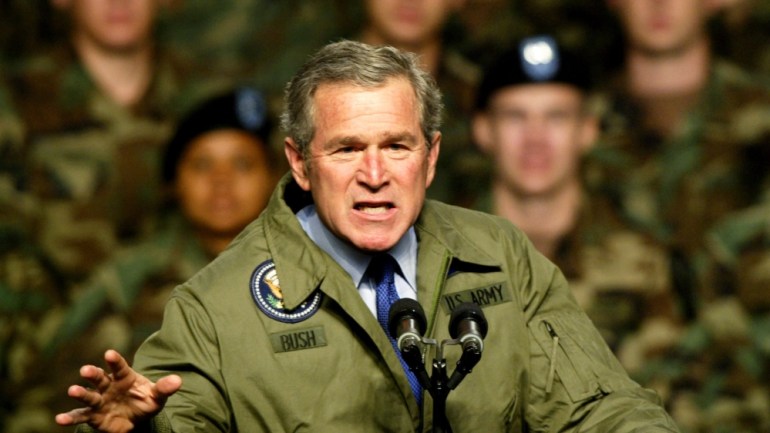 Bush in Army jacket that says "Bush" on one side and "US Army" on the other, at the microphone in the middle of a speech. His face is frozen in a fierce expression. Behind him, blurred, is a group of US soldiers.
