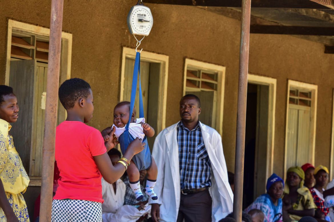 A mother weighs her baby at Kibibi Health center in Budondo Sub county Jinja District, as Musomba Joshua (R) watches, Musomba is an enrolled comprehensive Nurse at the facility.