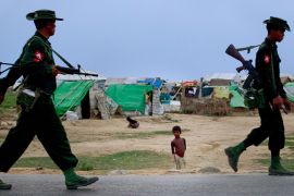 In this May 13'' 2013 photo, an internally displaced Rohingya boy, center watches army soldiers on foot-patrol in the foreground of makeshift tent camps for Rohingya people in Sittwe, northwestern Rakh