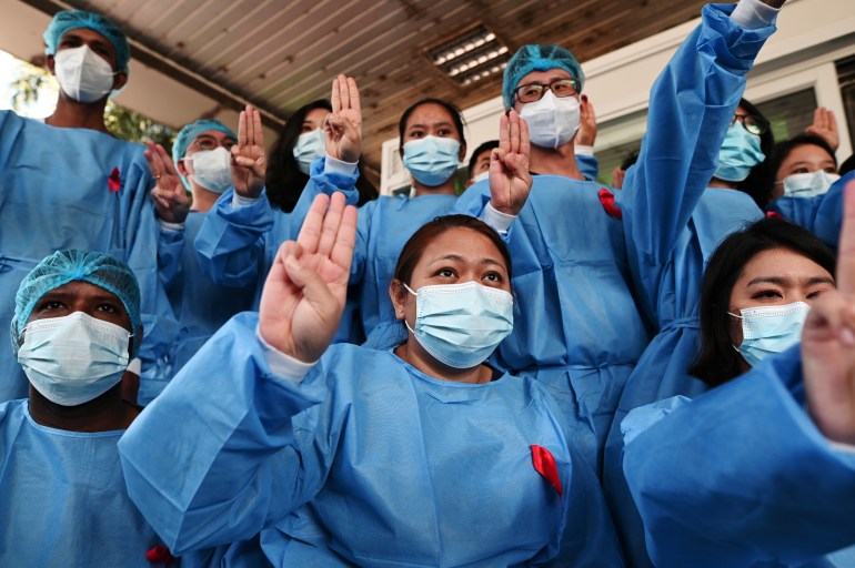 Health workers in blue medical gowns and face masks join mass protests against the military in Myanmar. The photo shows a group of workers, some women and some men, holding up the three finger salute.