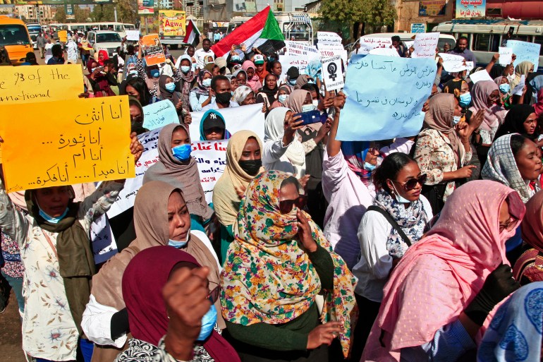 A large group of Sudanese women, many holding signs in Arabic, protesting in a street.