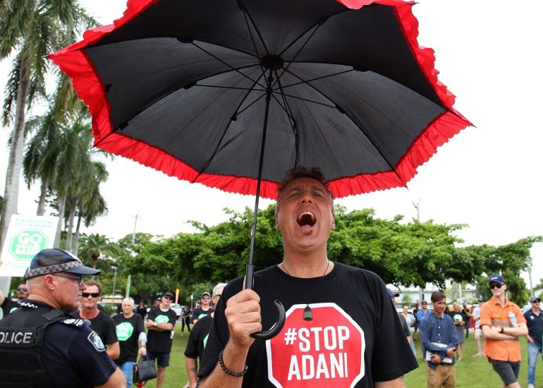 An activist at a rally in Mackay, Queensland in Australia, wearing a black T-shirt with "STOP ADANI" on it written in white letters in a red, STOP-sign logo. He is holding a black umbrella with red frilling going around it.