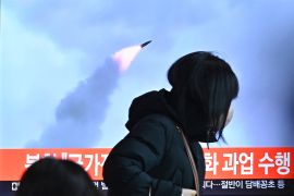 A woman in a heavy jacket and hat is silhouetted against a television screen showing a rocket launch in a report about Pyongynag;s second missile test in a week