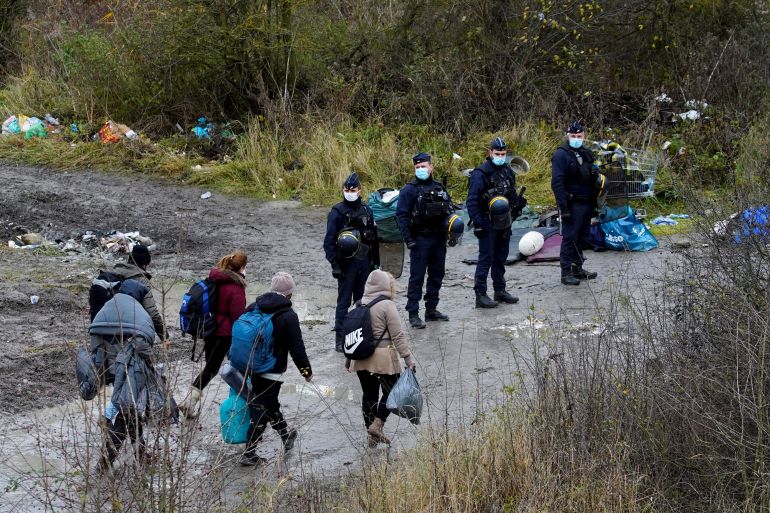 Migrants walk while police officers stand guard as they dismantle a makeshift migrant camp at Loon Beach, near Dunkirk, France