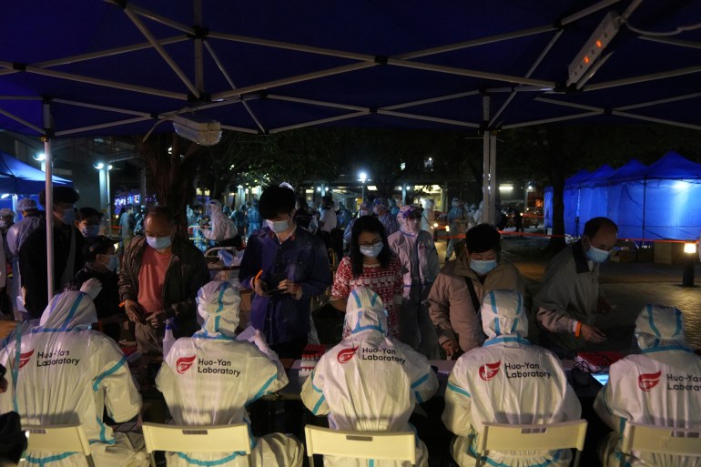 A row of health workers in hazmat suits sit on a bench, their backs to the camera - as residents of Kwai Chung queue up in the dark for COVID tests