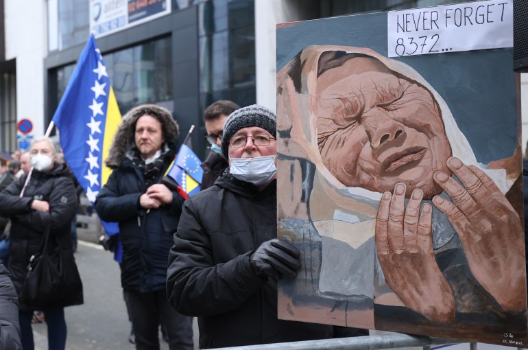 A woman at a protest in Brussels holds up a painting showing a mother from Srebrenica crying with the message "Never forget 8,372..." written at the top