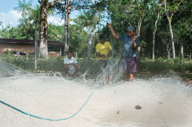 Elder Júnior is a man wearning a blue t-shirt. He stands and mends a fishing net 