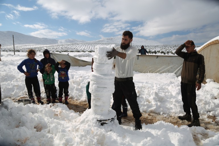 A man builds a snowman as children watch on in Sheikh Bilal Camp in Afrin. [Courtesy of Ali Haj Suleiman]