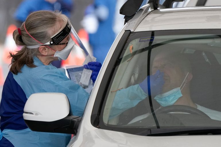 A woman in protective clothing administers a COVID test to someone sitting in a car