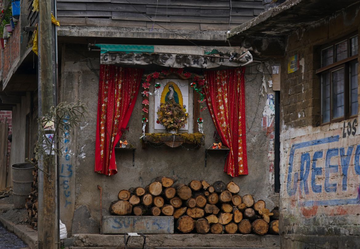 A Virgin of Guadalupe altar decorates the facade of a home where firewood used for cooking is piled below
