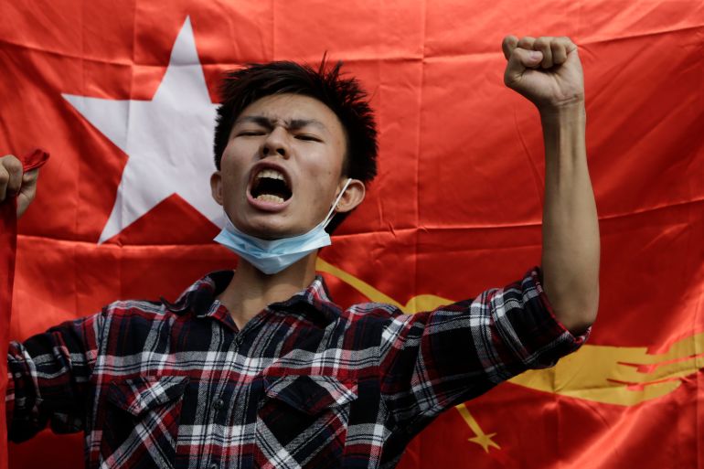 A young man in a red checked shirt shouts slogans and raises his arm in the air in front of the NLD's red flag
