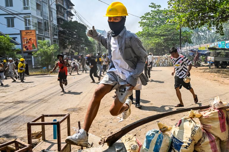 A protester in a yellow ghard hat jumps over a makeshift barricade as he flees security forces in Yangon in March