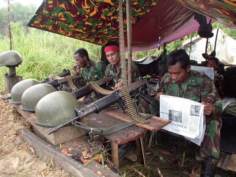 Indonesian soliders sitting under a shelter with guns and helmets on a makeshift table in front of them, in Aceh province