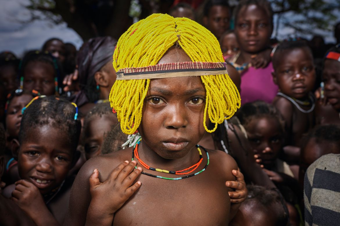A photo of a refugee girl wearing a traditional beaded wigs called Ena meant for girls of the Mudimba tribe from southern Angola with a group d children in the back.