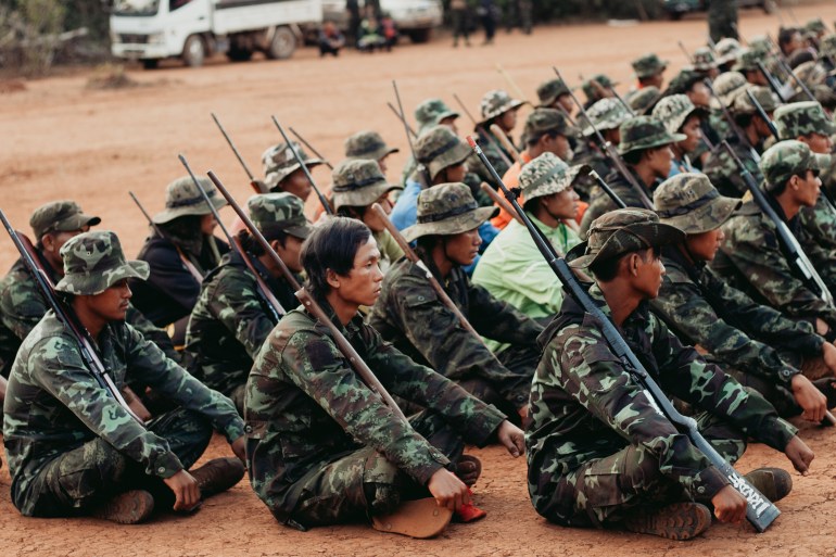 A group of fighters sitting on the ground in a training camp, holding rifles.