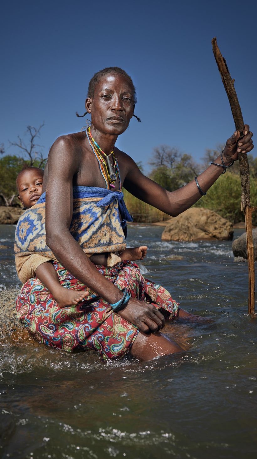 A photo of a woman with a child strapped to her back as they cross the river to get to Namibia from Angola.