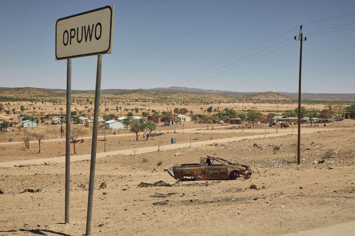 A photo of an empty road with a rusty car and a sign that says “OPUWO”.