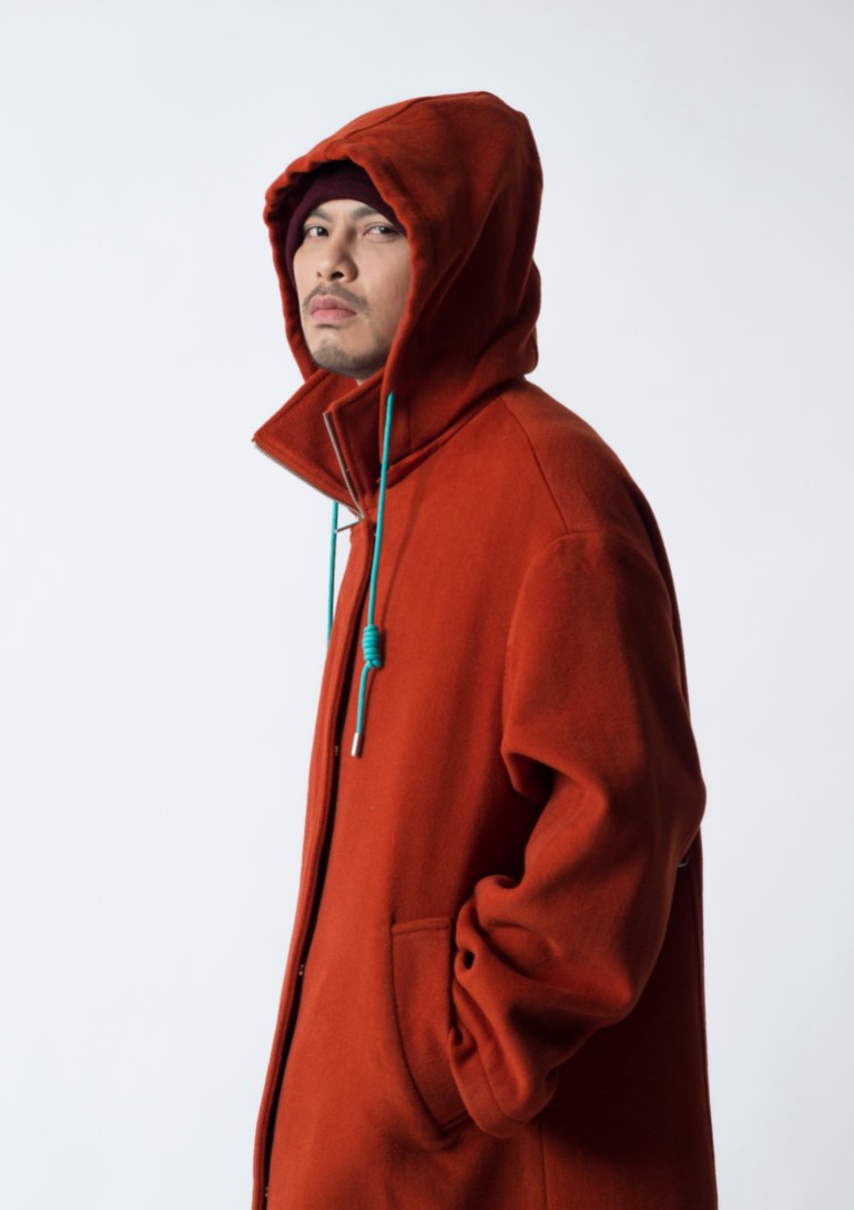 A publicity photo of Malaysian performer Namewee dressed in a red hoodie with the hood pulled up and his hands in his pockets