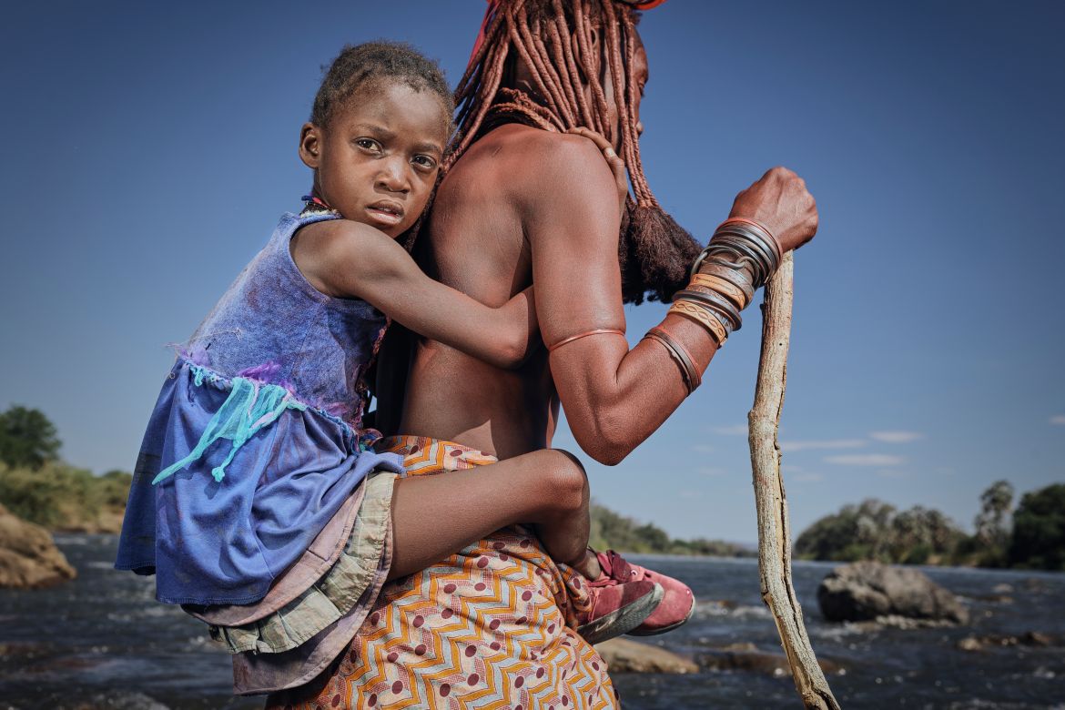 A photo of a woman holding a cane, carrying a child on her back.