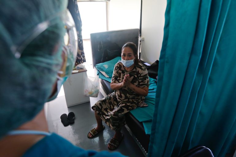 A woman sits on a bed behind blue curtains in a COVID isolation ward as a medic in protective clothing looks in