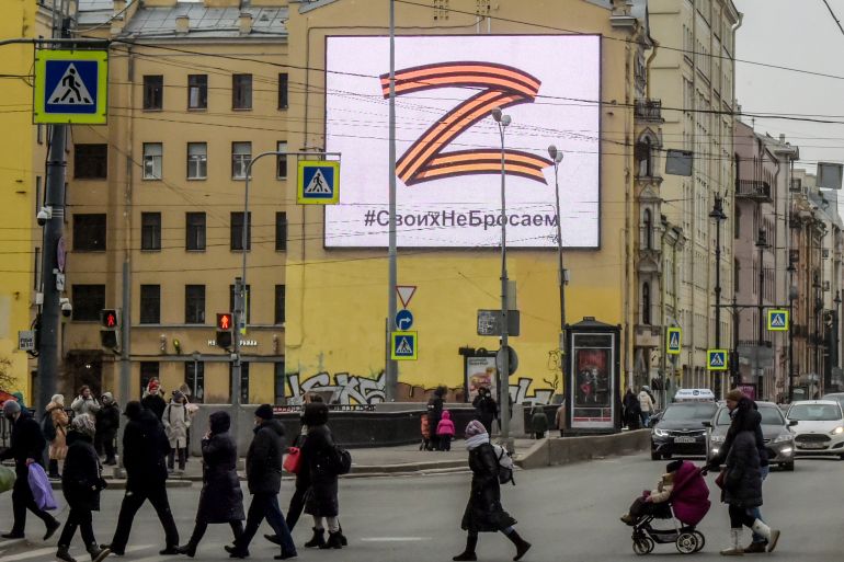 Pedestrians cross a street in front of a billboard displaying the symbol "Z" in the colours of the ribbon of Saint George and a slogan reading: "We don't give up on our people", in support of the Russian armed forces, in Saint Petersburg