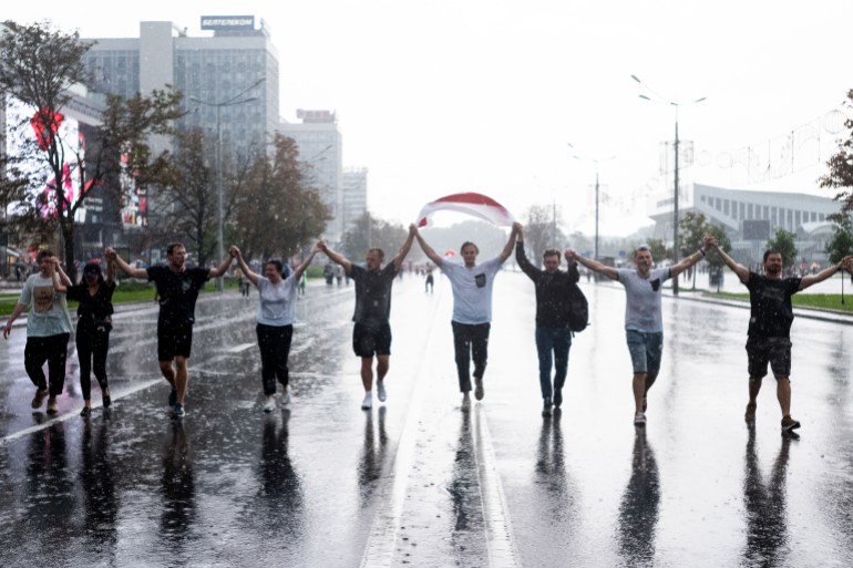 A photo of nine people holding hands and walking in the middle of the street with a Belarusian flag.