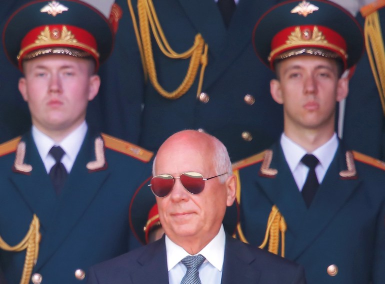 Rostec CEO Sergei Chemezov in mirrored glasses standing in front of two soldiers in military uniform