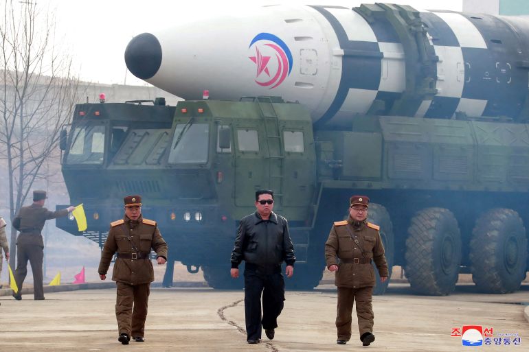 Kim Jong Un in black leather jacket and sunglasses and flanked by two men in military uniform walking away from a white-coned missile on its transporter