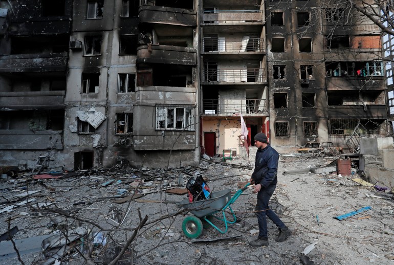 A resident pushes a wheelbarrow in front of a destroyed building in Mariupol