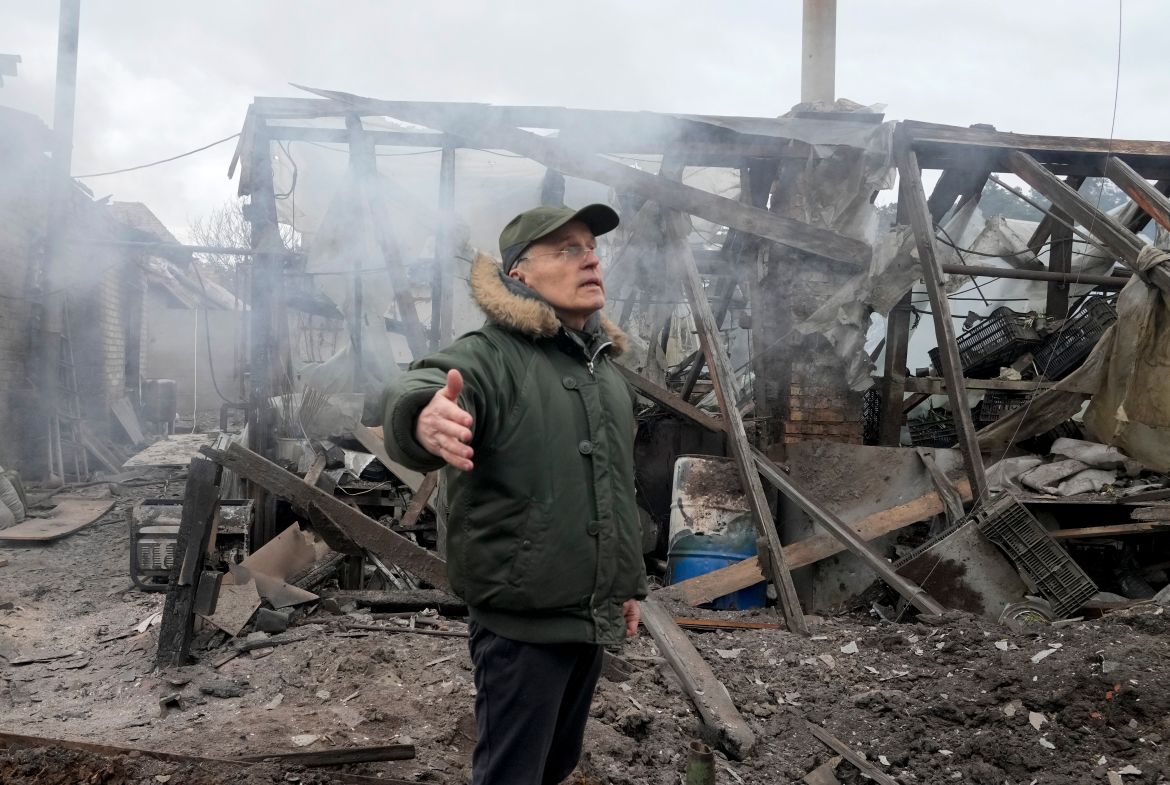 A man opens his arms as he stands near a house destroyed in the Russian artillery shelling