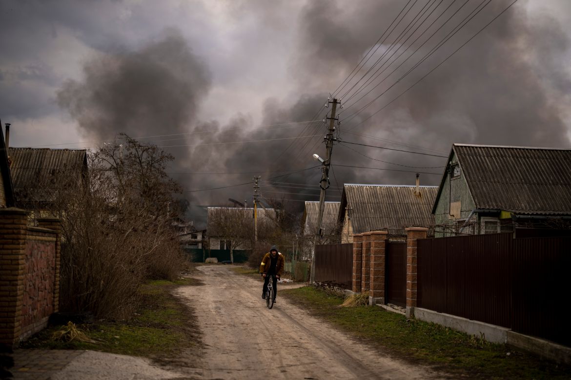A Ukrainian man rides his bicycle near a factory and a store burning after they had been bombarded in Irpin