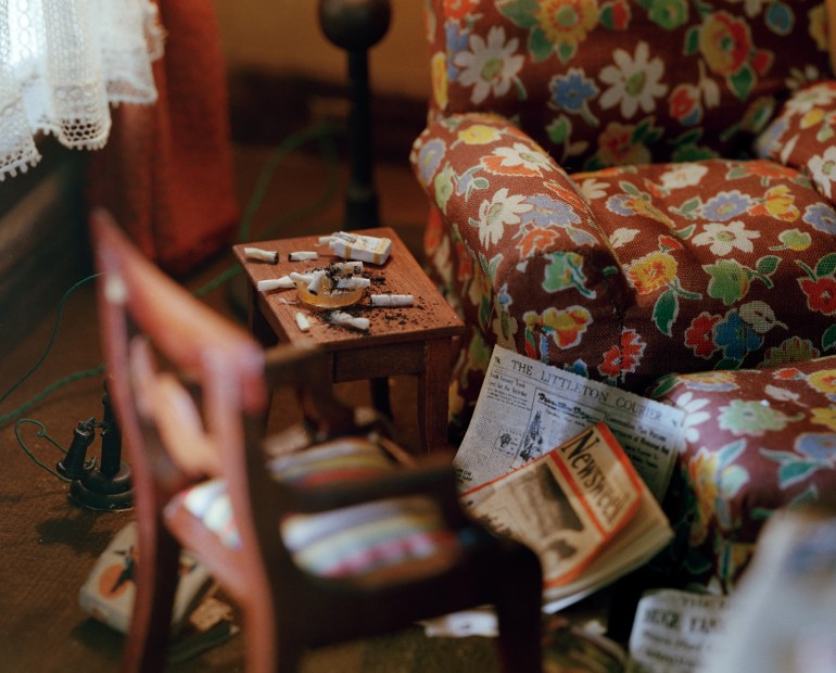 A photo of a diorama of a living room with cigarettes on the side table to the sofa with a chair and newspapers and magazines on the floor.