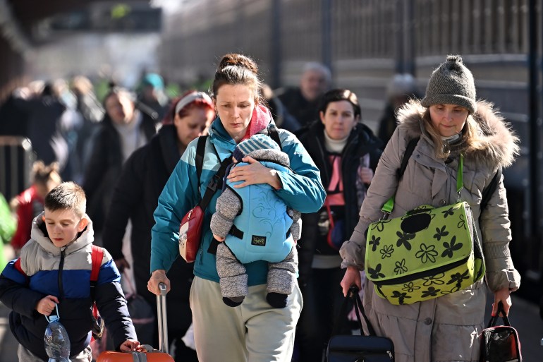 A photo of people walking with suitcases and bags with three people in focus, a child on the left, a woman in the middle holding a baby and a woman on the right.