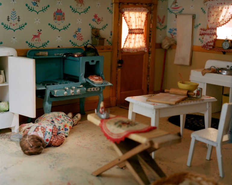 A photo of a diorama of a doll lying on it's back in the kitchen in front of the stove.