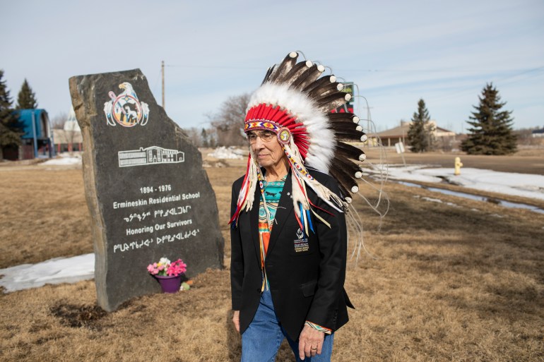 A photo of Chief Willie Littlechild walking past a plaque marking the Ermineskin Residential School in Maskwacis, Alberta that says "1894-1975 Ermineskin Residential School, Honoring our survivors"