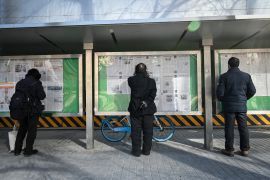 Three men standing on the street to read Chinese state-run newspapers that have been pinned up on glass-fronted boards.