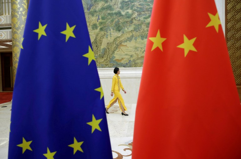 Woman in yellow suit walks through a foyer, in the distance, in between a close-up of flags of China and the European Union.