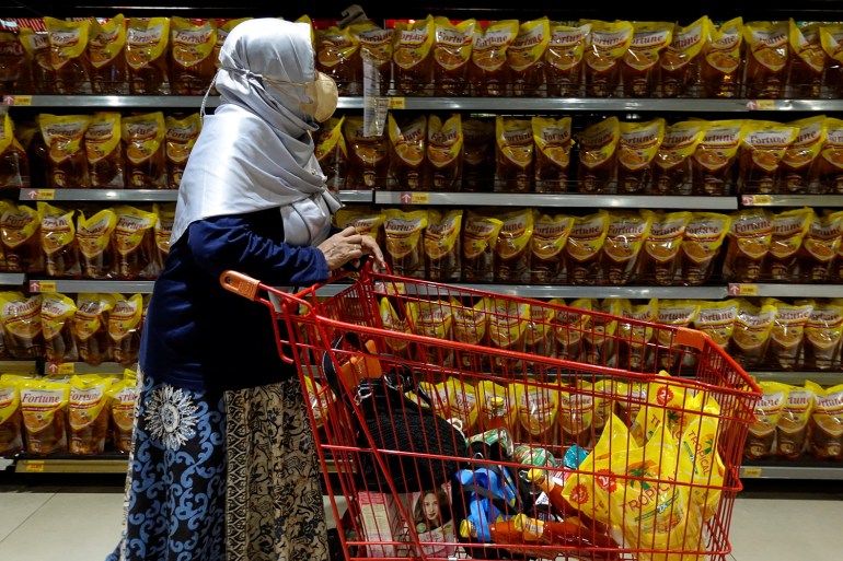 A woman in Jakarta in a shiny, white headscarf, pushes her trolley down a supermarket aisle stocked with cooking oil made from palm oil