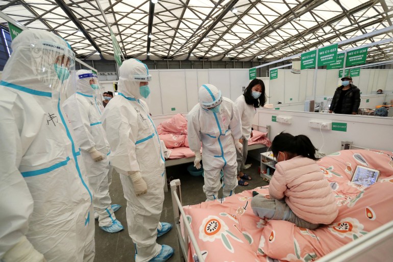 A young girl in a pink puffer jacket sits on her bed as hazmat suited officials inspect a mass quarantine centre in Shanghai, China.