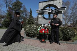 A Russian orthodox priest in black robes with a Black Sea Fleet veteran at a ceremony in honour of the Moskva in front of a Soviet-style sculpture strewn with flowers