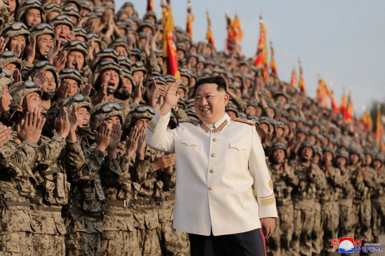 North Korean leader Kim Jong Un in white uniform waves and smiles as he walks past a group of cheering soldiers in a military parade.