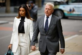 Boris Becker in a grey suit and tie walks to court with his partner partner Lilian de Carvalho Monteiro in white trouser suit and black shirt