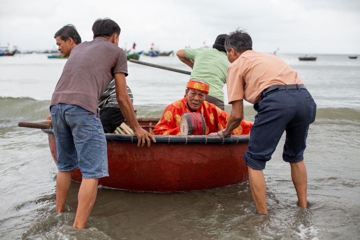 A photo of fishermen ferrying an elderly whale worshipper in crimson-and-gold áo dài.