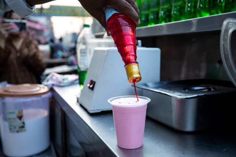 A hand is seen swirling straight rooh afza onto the top of a cup of milk blended with more rooh afza