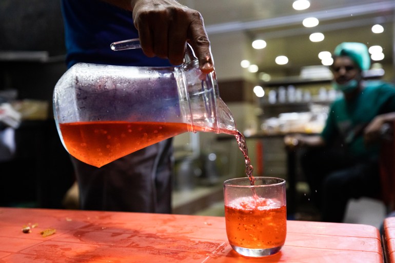 A hand is pouting prepared (diluted) rooh afza out of a glass pitcher into a short glass. Another person can be seen in the background, out of focus