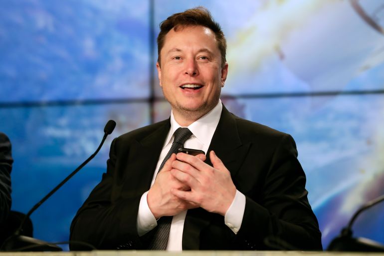 Elon Musk smiles from behind a microphone while holding his mobile phone near his chest.